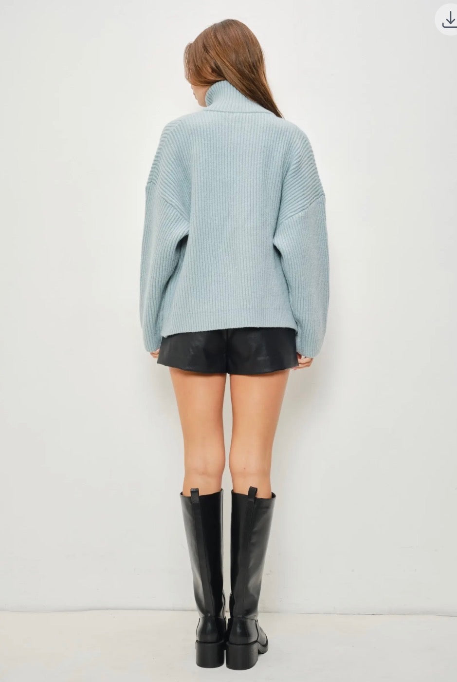 A woman shown from the back wearing a light blue ribbed knit mock-neck sweater, black leather shorts, and black knee-high boots against a plain white wall.