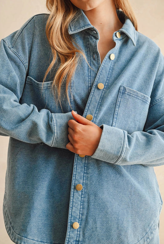 A close-up of a person wearing an oversized light blue denim button-up shirt with gold-tone buttons, shown against a plain beige background.