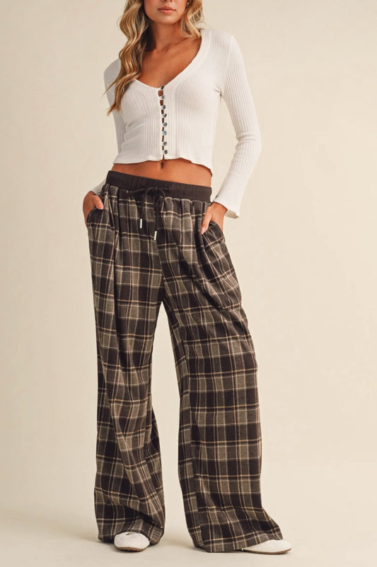 A model wearing a white ribbed long-sleeve crop top with front buttons and wide-leg plaid pants in brown, beige, and black, standing against a solid beige studio background.